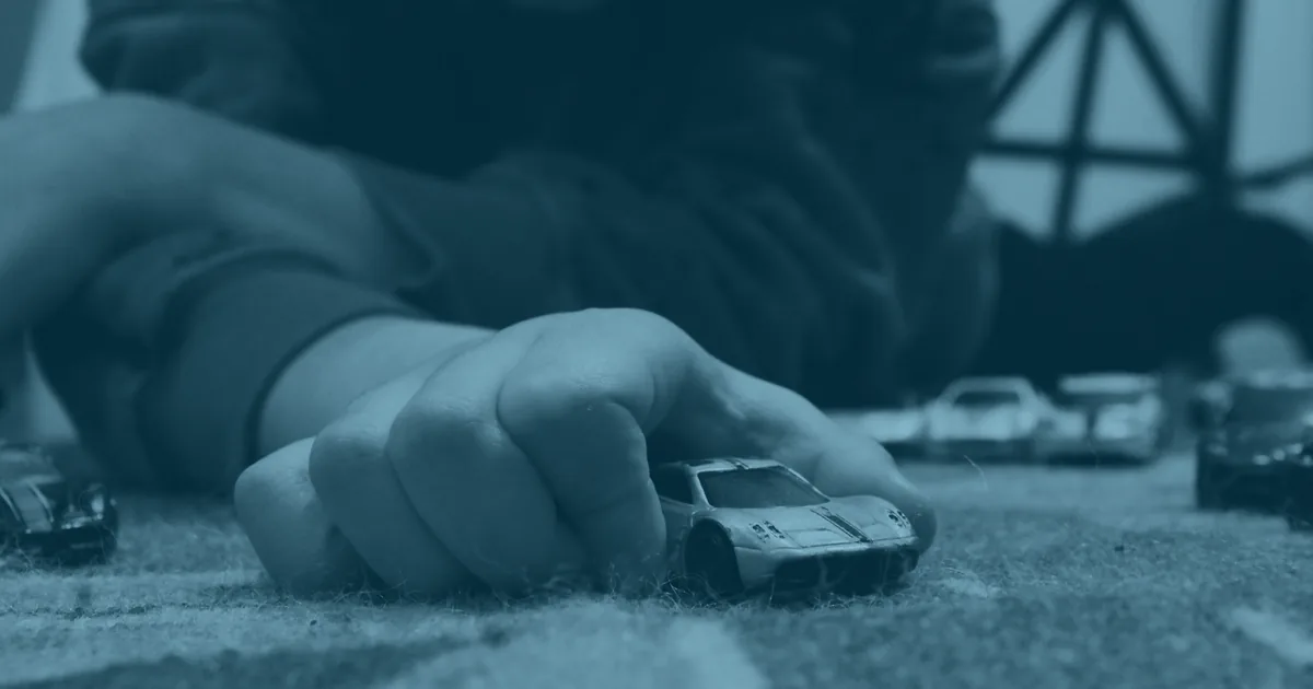 A child lying on a patterned rug, pushing a small toy car along with one hand while more toy cars sit scattered around.