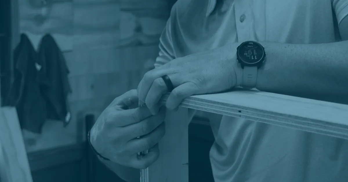 A man in a workshop steadying a panel of plywood with both hands, a wedding ring on his finger and a sports watch on his wrist.