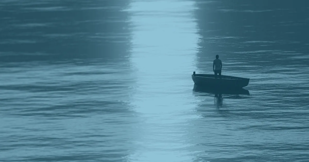 A lone figure standing upright in a small boat far out on the water, with the low sun laying a broad band of light across the surface.