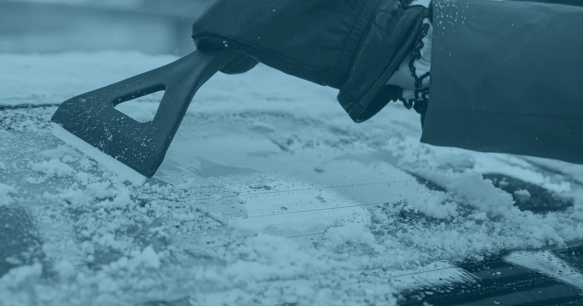 A gloved hand scraping a thick layer of ice and snow off a car windscreen.