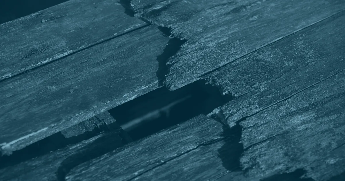 Weathered decking boards split and rotted through, with a gap opening between them in low raking sunlight.