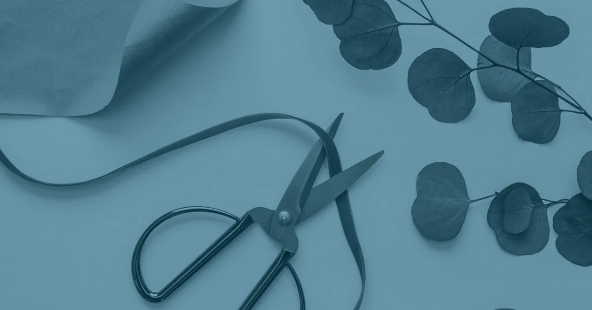 Wrapping materials laid out on a table: a pair of scissors, a curled length of ribbon, a folded sheet of kraft paper and sprigs of eucalyptus.